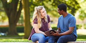 A couple of college student sitting together outdoors talking and looking at an iPad.