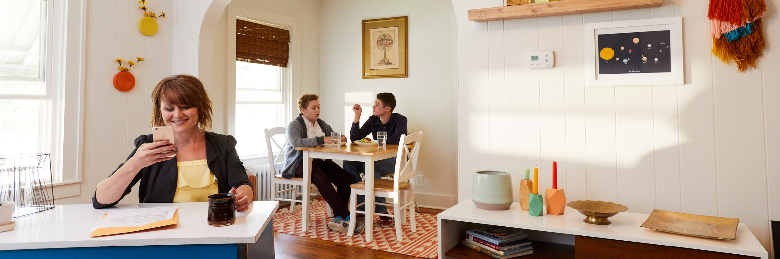 A woman looks at her phone while sitting at a table, with two boys sitting behind her at another table.