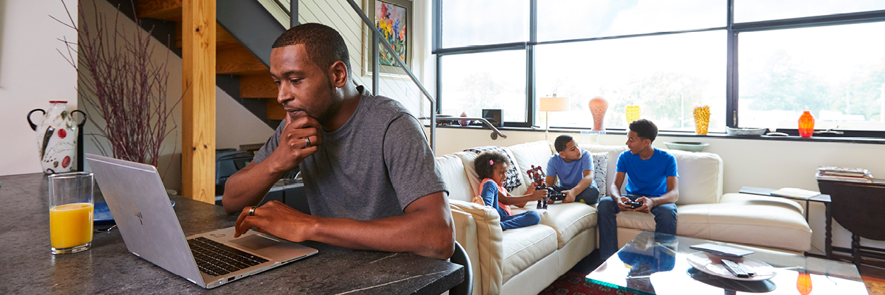 A man sit at a table looking at his laptop. Three children--two boys and a girl--sit on a couch behind him, talking to each other.