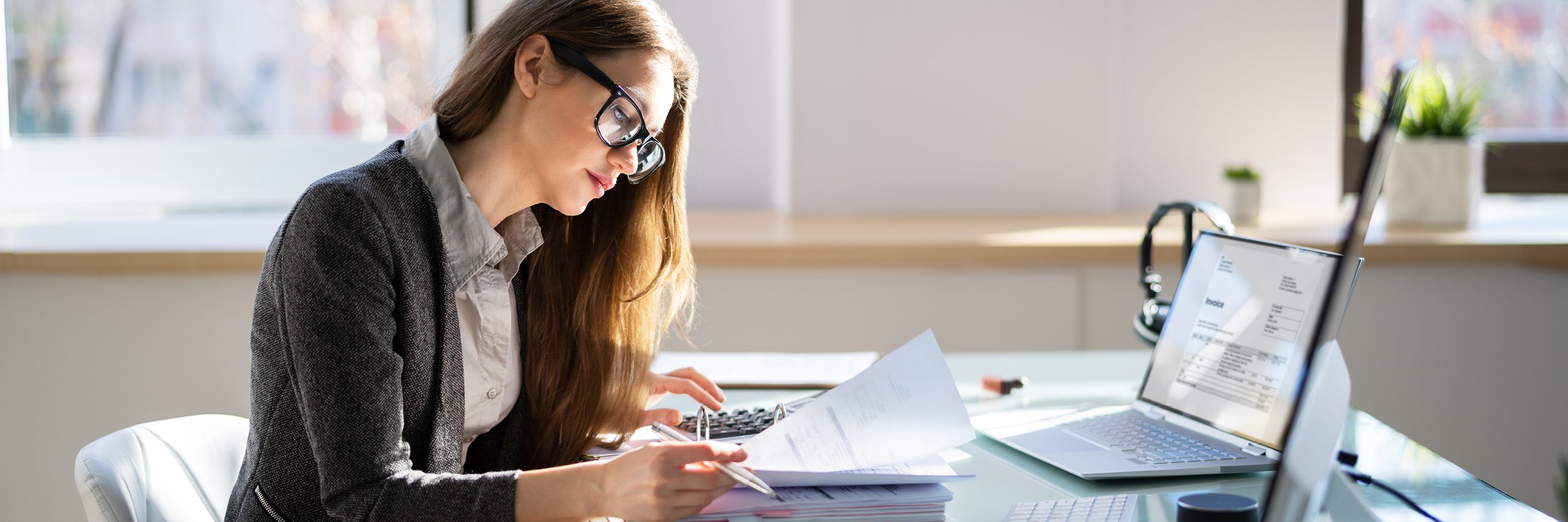 A woman sits at a desk, looking at some papers and has her laptop open.