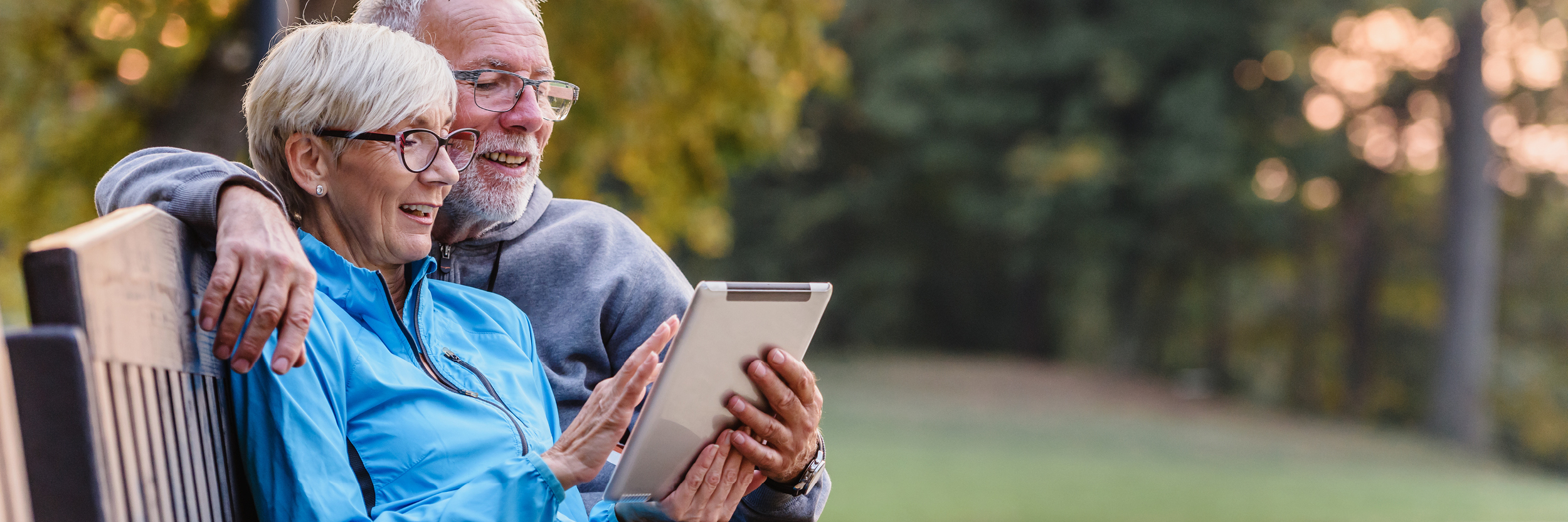 A mature man and woman sit on a park bench looking at an iPad.