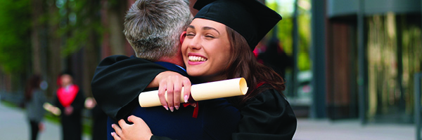 A woman wearing a graduation gown and holding a diploma hugs a man