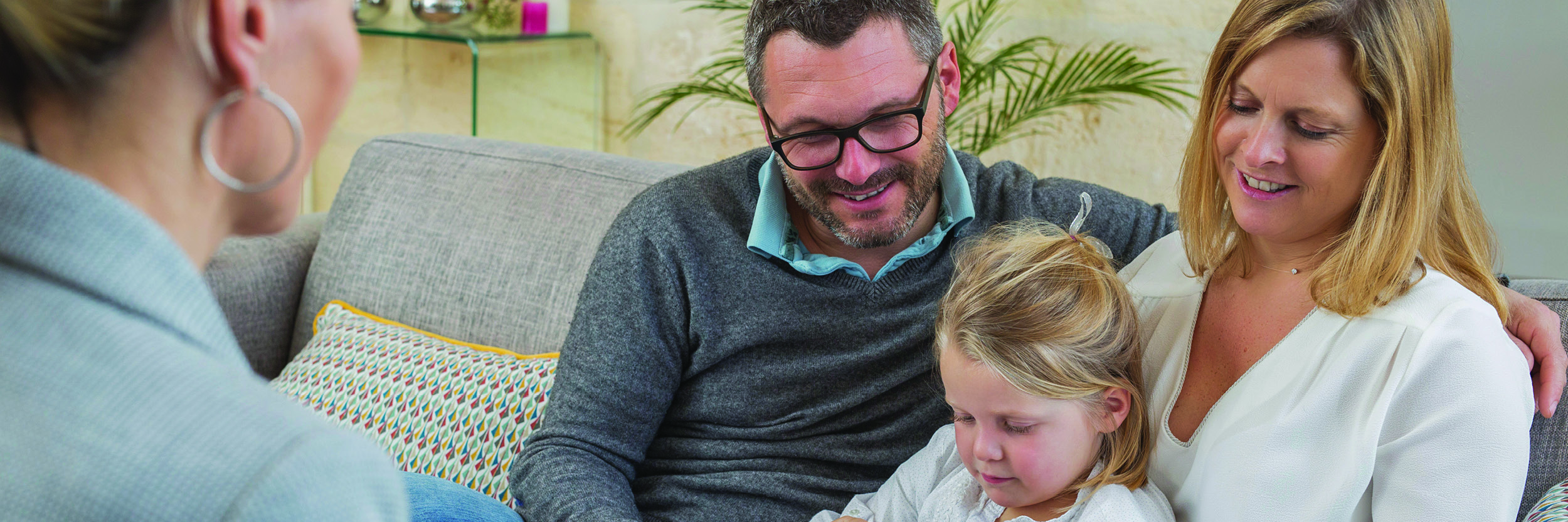 A man and woman are sitting on a couch looking at paperwork with a little girl between them. Another woman is facing them but only her back is seen.