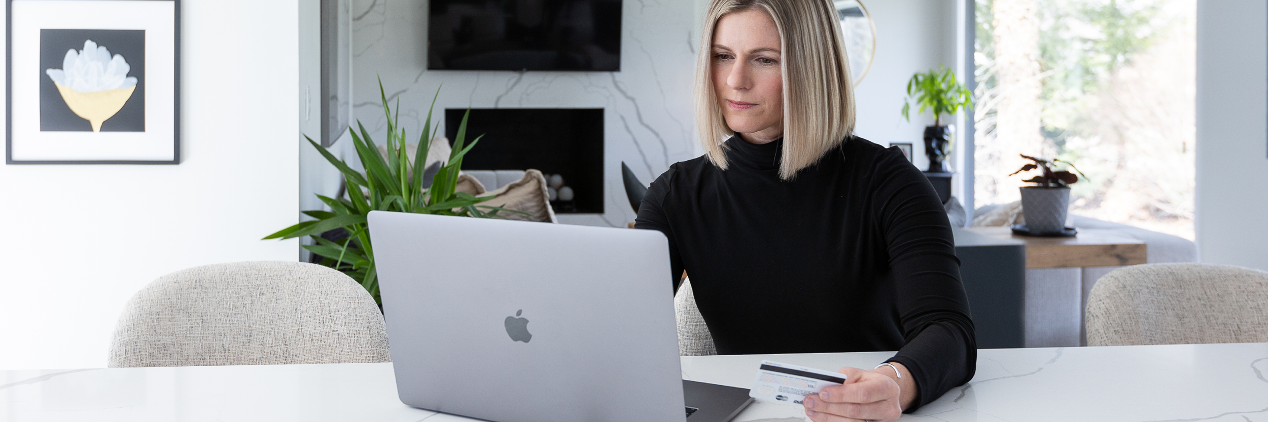 A woman holds a credit card while she sits at a table, looking at a laptop.