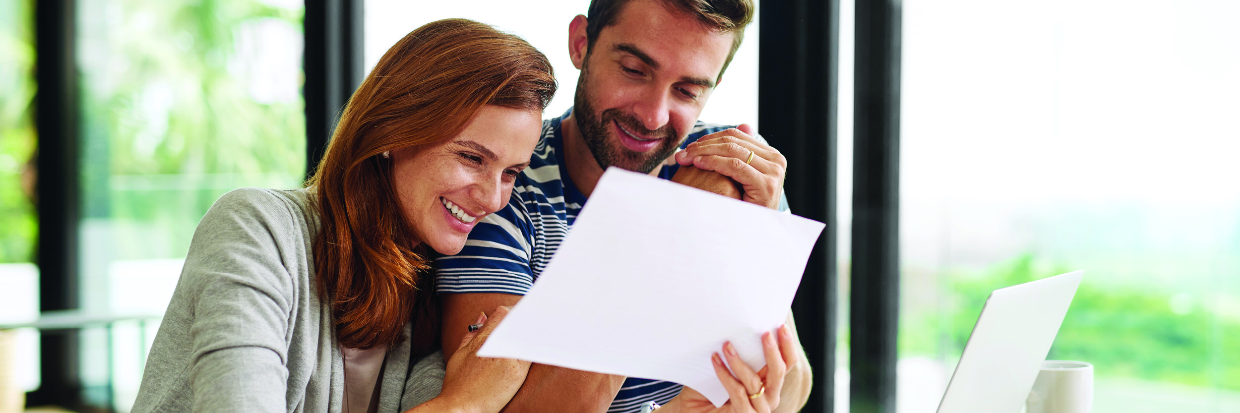 A man and a woman sit at a table. The woman has her arms around the man and they're looking at a piece of paper.