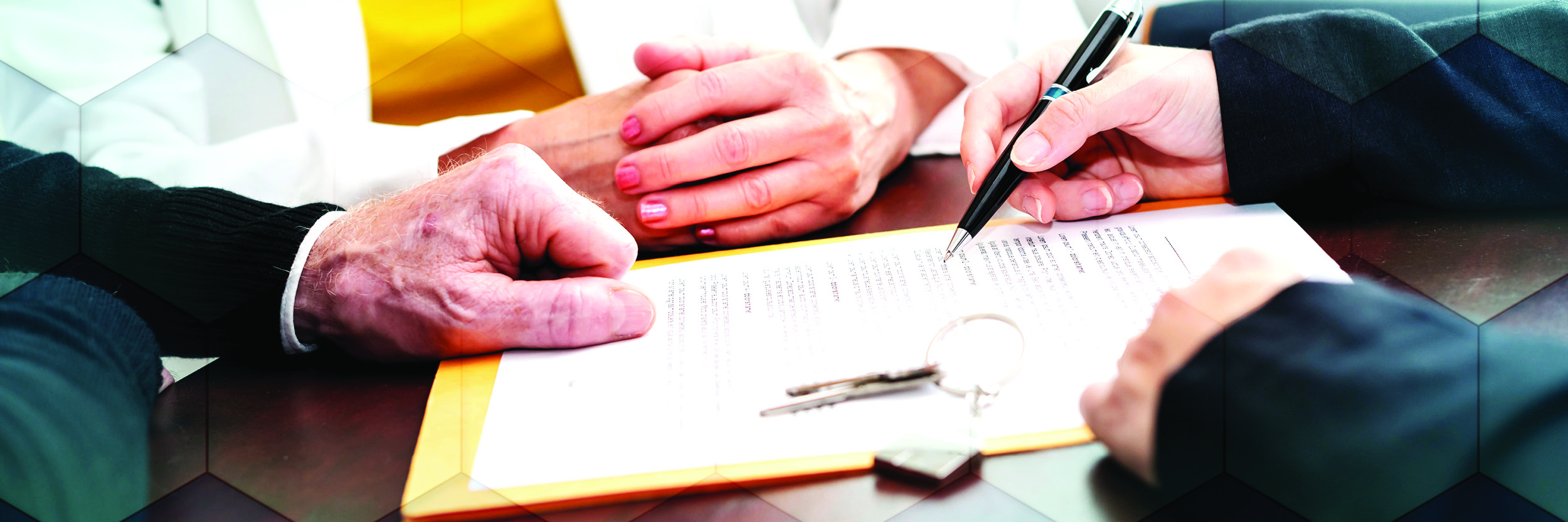 The hands of three people are seen as they sit around a table. One is holding a pen and a piece of paper is on the table.