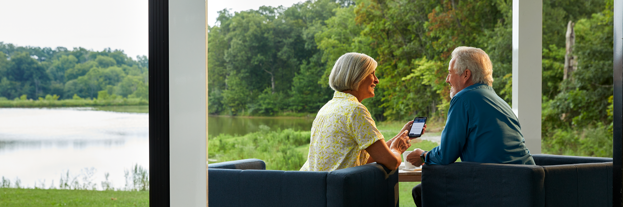 A man and woman sit on a porch with a lake view as they look at a cell phone.