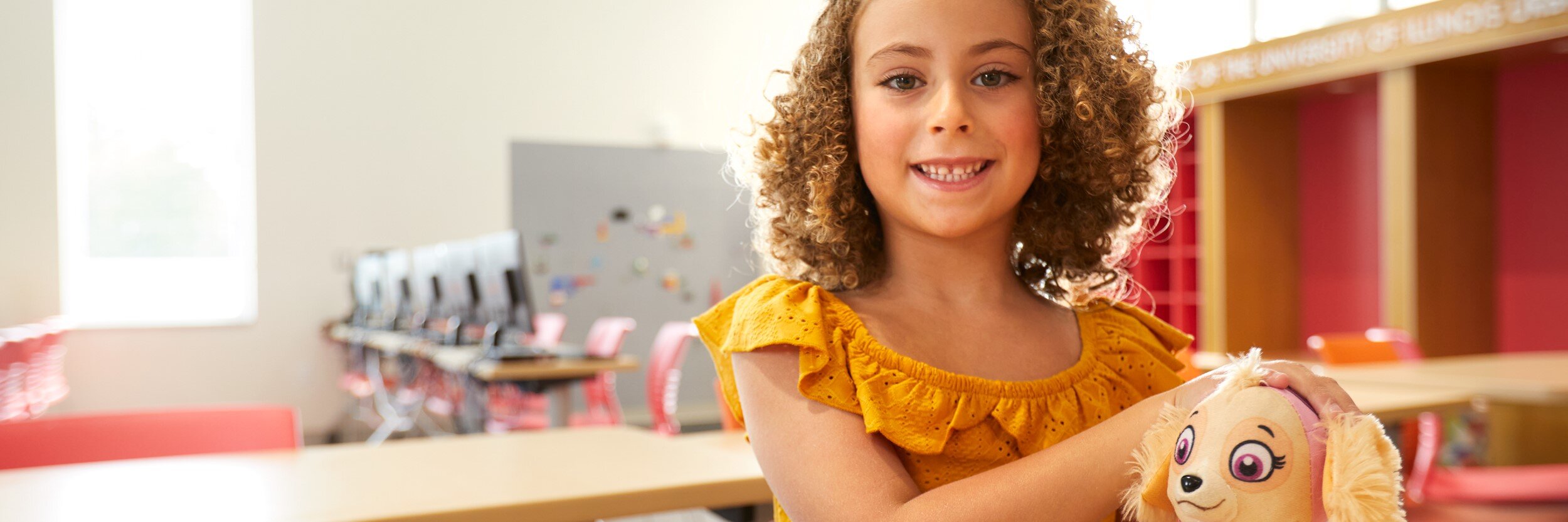 A young girl smiles as she holds a stuffed animal in a classroom.