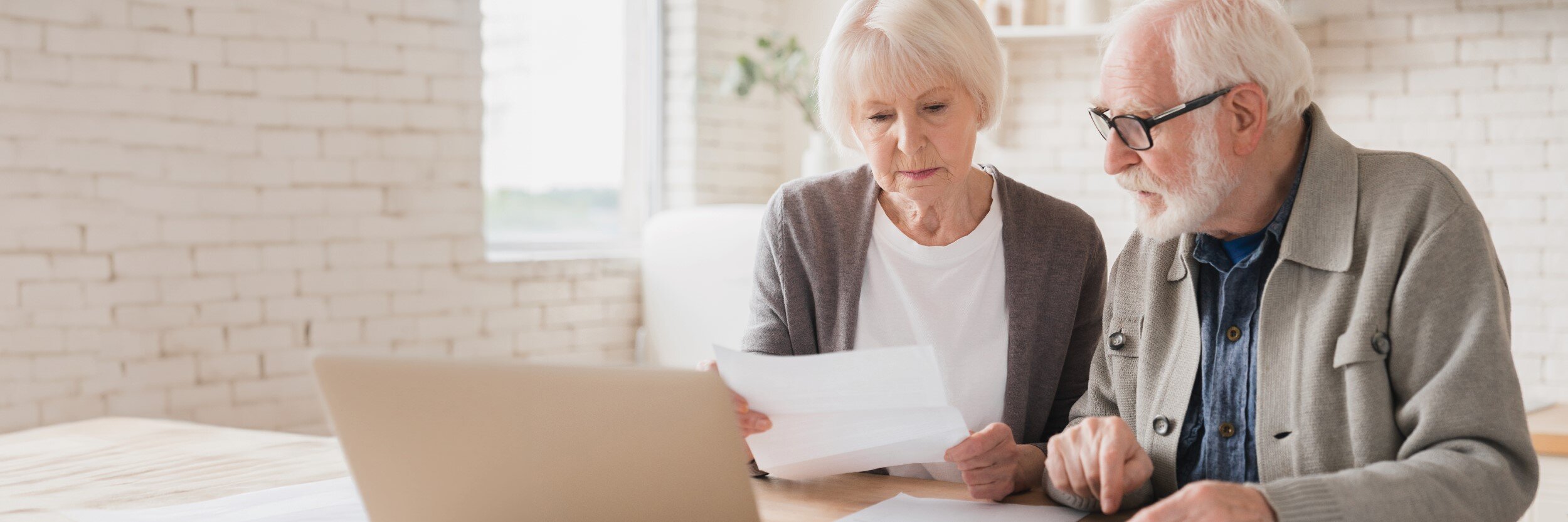 A mature man and woman sit at a table with a laptop open as they look at a piece of paper.