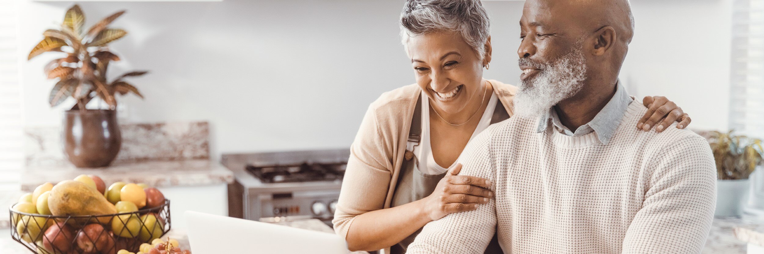 An older man and woman are at a kitchen counter looking at a laptop, while they laugh.