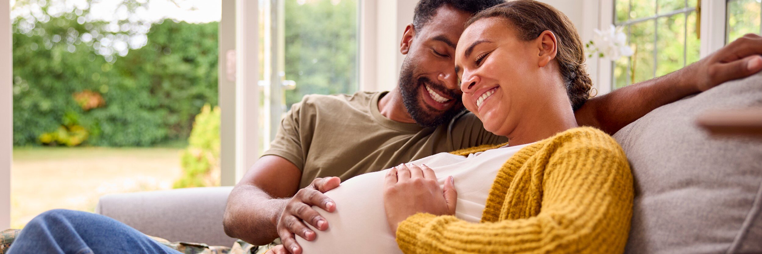 A man and a pregnant woman sit on a couch, smiling.
