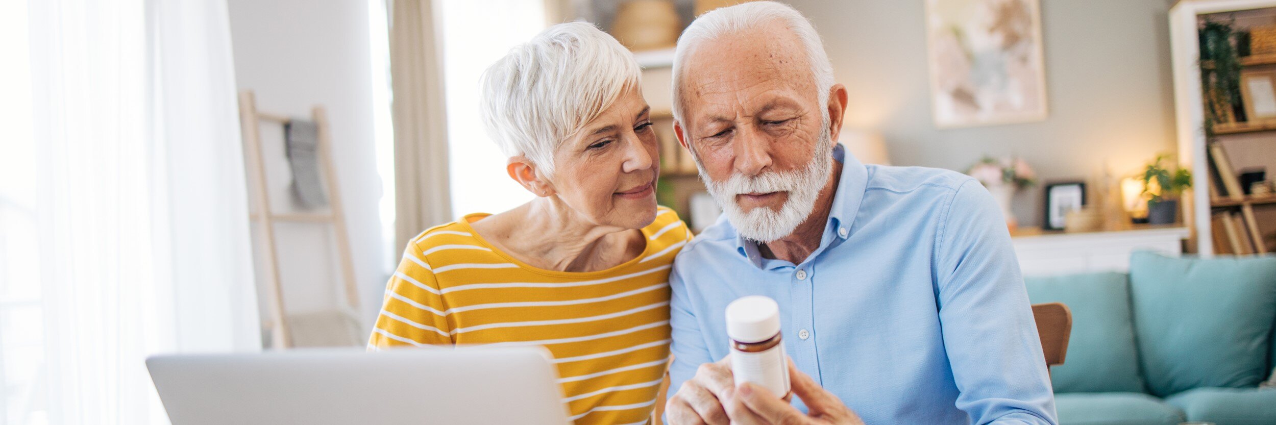 A man and woman sit in front of a laptop, looking at a prescription bottle.