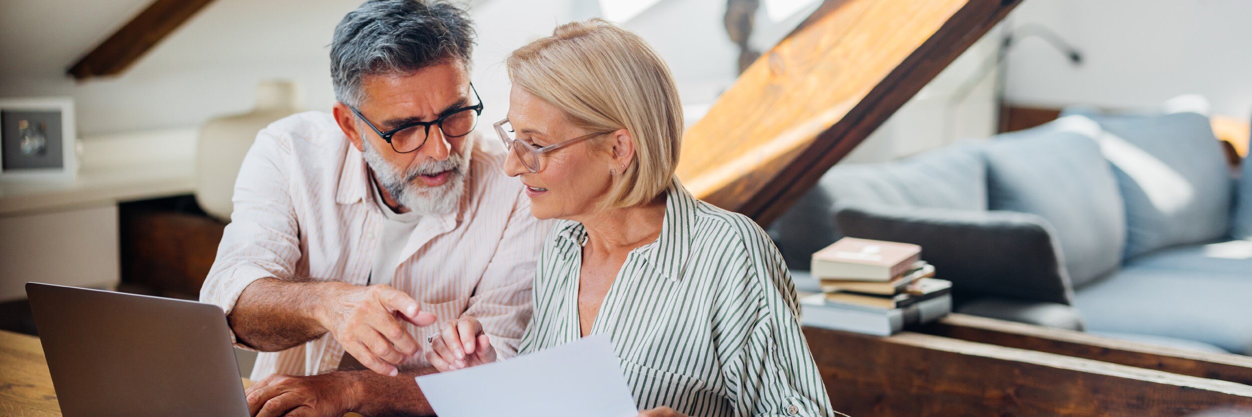 A man and a woman sit at a laptop, holding paperwork.