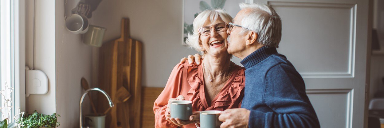 An older man kisses an older woman on the cheek, while she laughs.