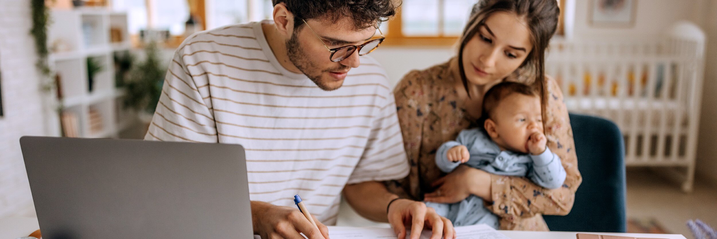 A man and a woman sit at a table with a laptop. A baby is on the woman's lap.
