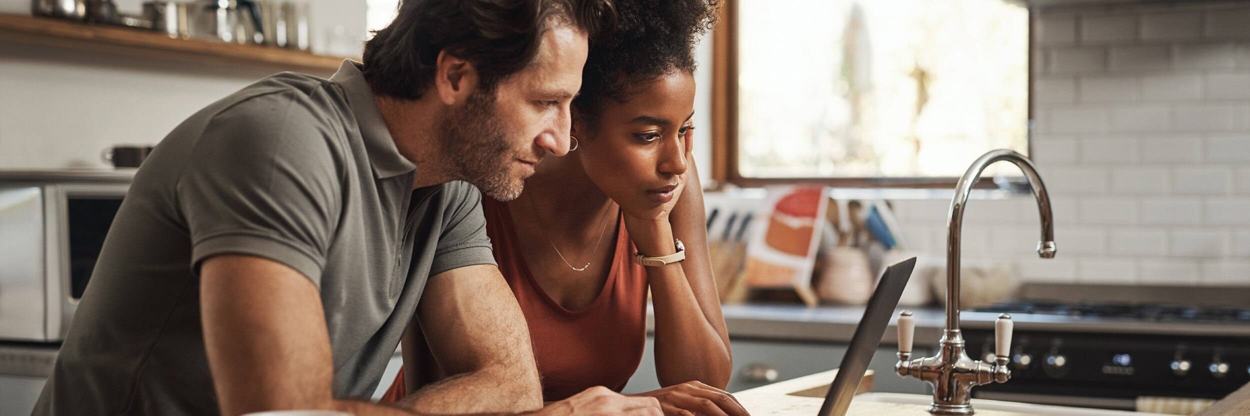 A man and woman sit at a kitchen counter looking at a  laptop