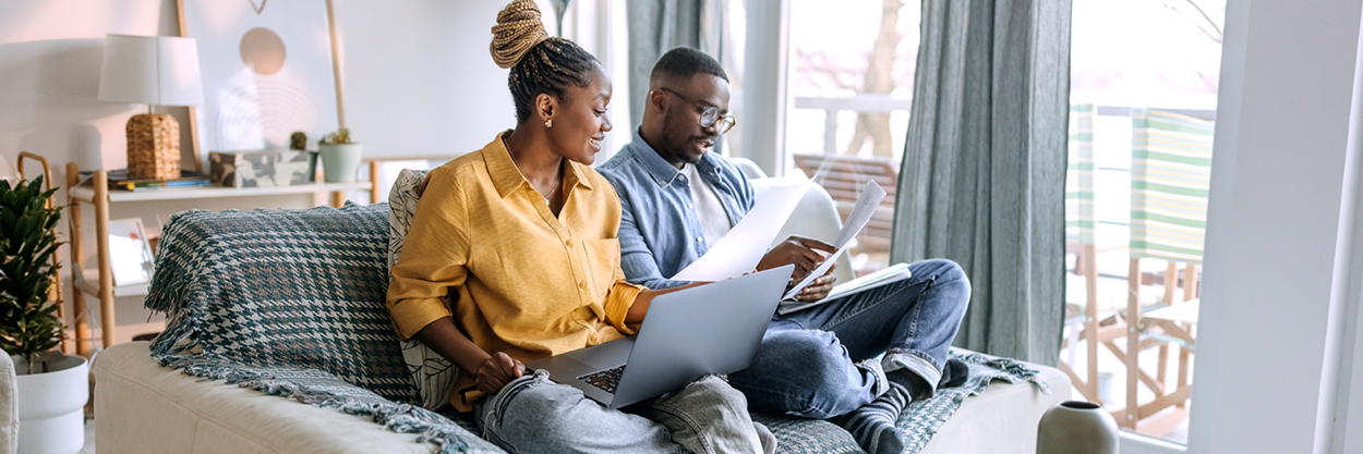A man and woman sit on a couch, looking at a laptop and paperwork.