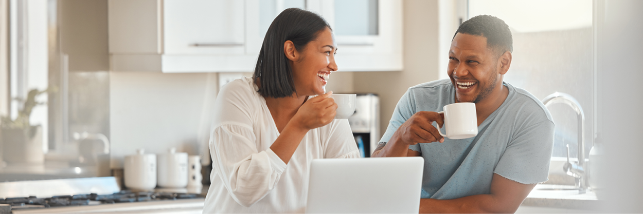 A man and woman sit at a kitchen counter, drinking coffee and looking at a laptop