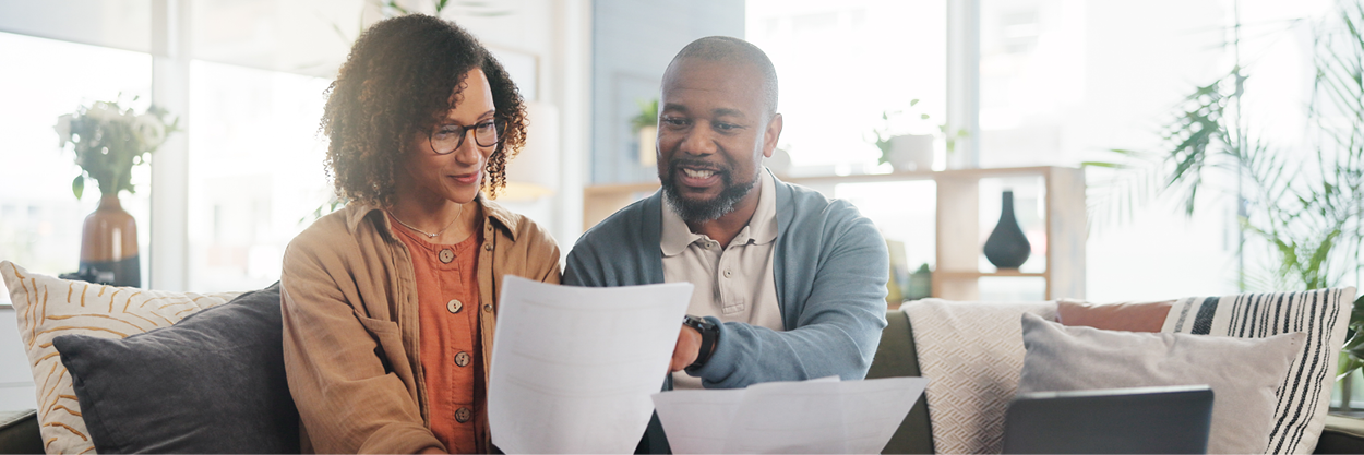 A man and woman sit on a couch, looking at paperwork.