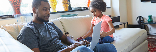 A man sits on a couch looking at his laptop while a little girl plays with a doll beside him.