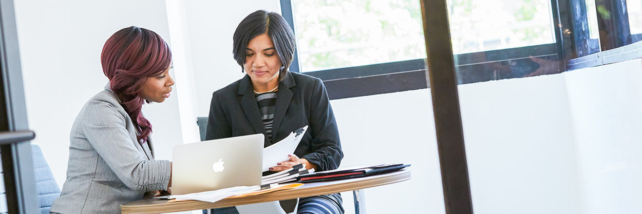 Two women sit at a desk looking at papers.