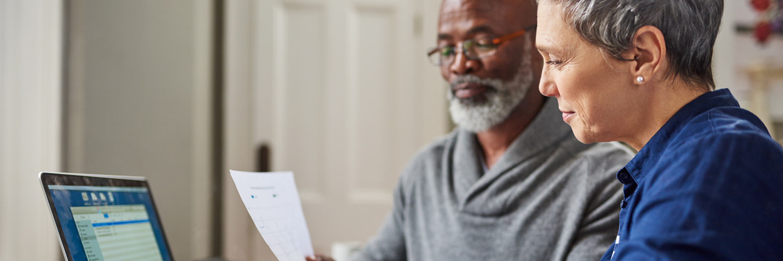 A man and woman look at papers while sitting before a laptop.