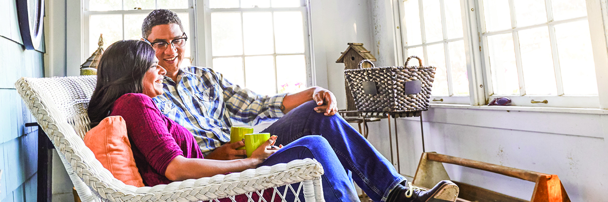 A man and woman sit on a wicker couch on a porch, holding cups of coffee, and talking.