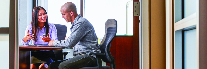 A man and a woman sit at an office table, talking.