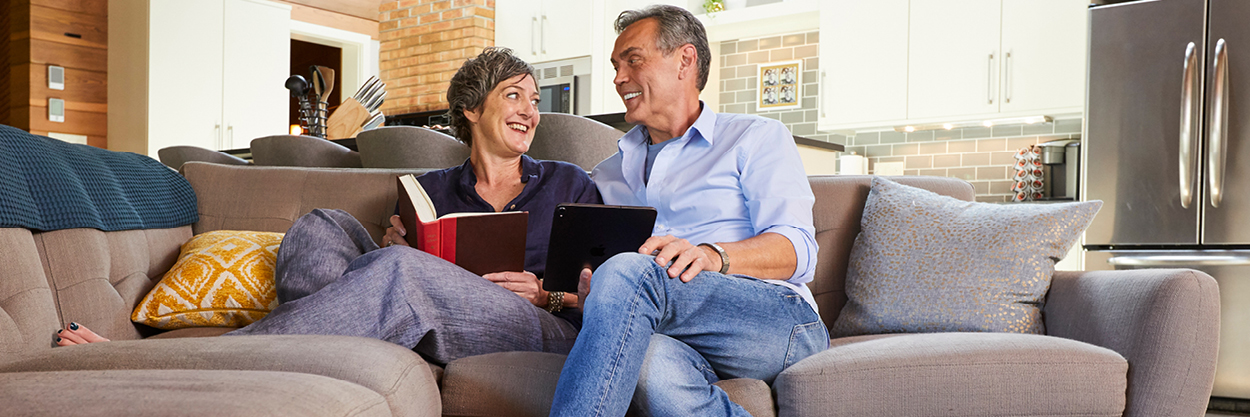 A man and woman sit next to each on a couch. The woman is holding a book and the man is holding an iPad. They are looking at each other and smiling.