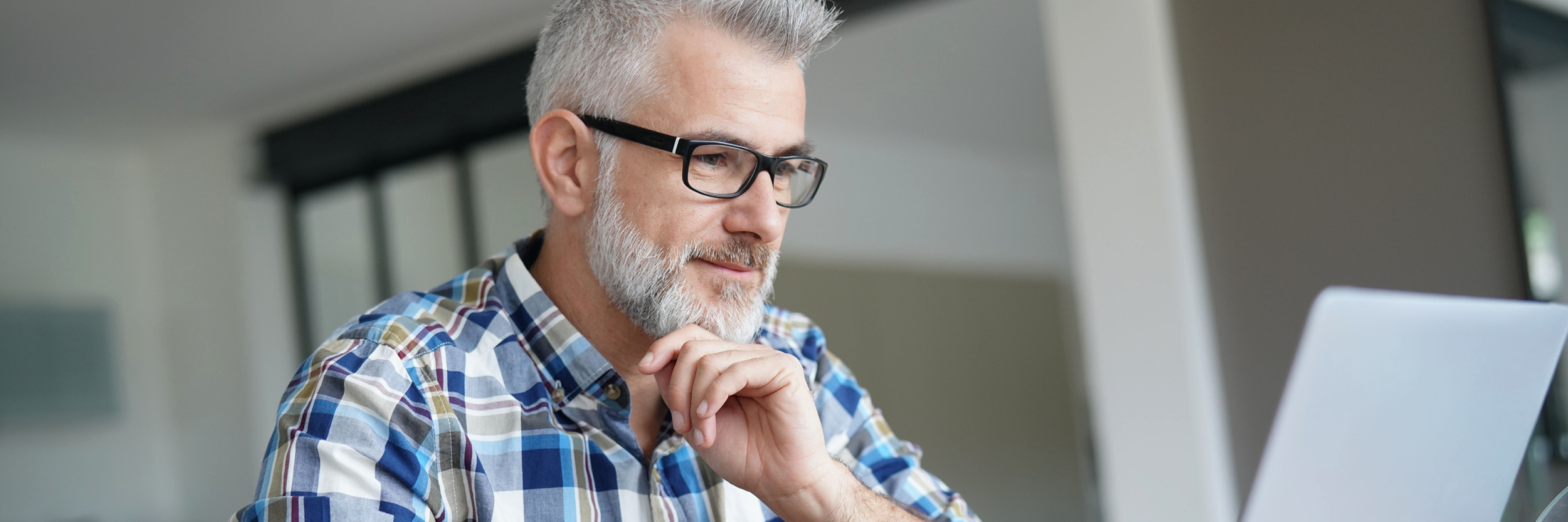 A man with a gray beard sits at a table, looking at a laptop.