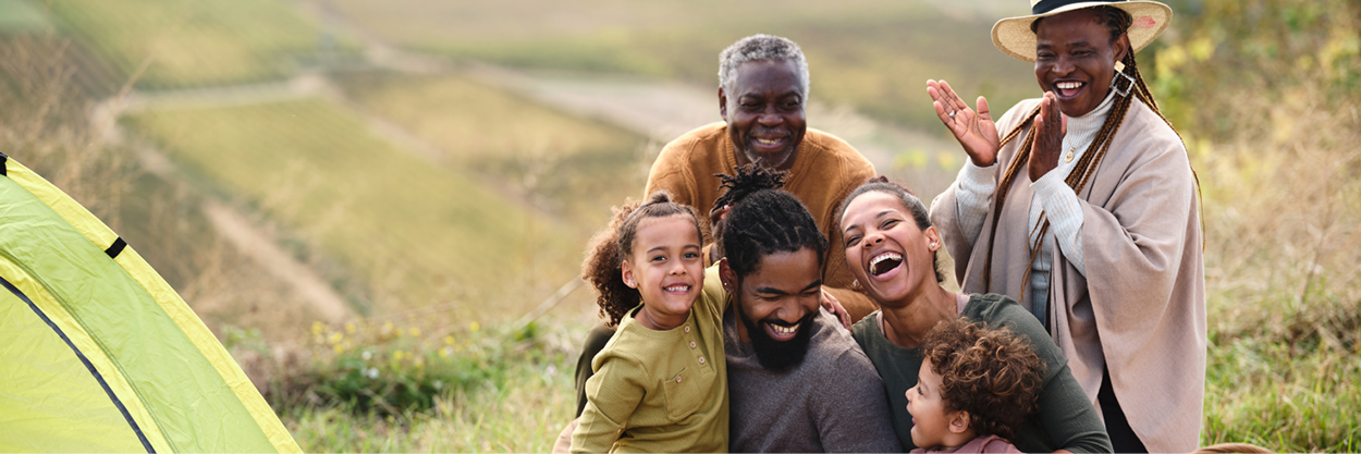A younger man and woman with two children and an older man and woman sit in a field near a tent.
