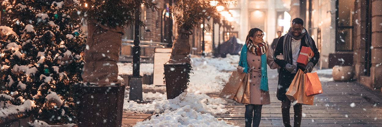 A man and woman walk in the snow, holding shopping bags