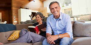 Man and woman sitting on couch, man smiling at camera while the woman reads a book.