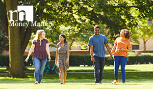 Four college students walking across the lawn on campus chatting and laughing together. 