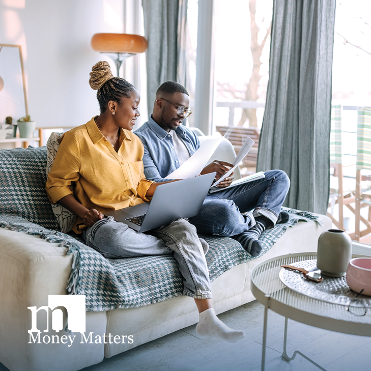 A man and woman sit on a couch, looking at a laptop and paperwork.