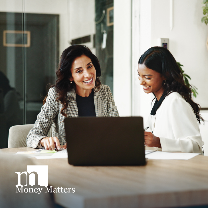 Two women sit at a table, looking at a laptop.