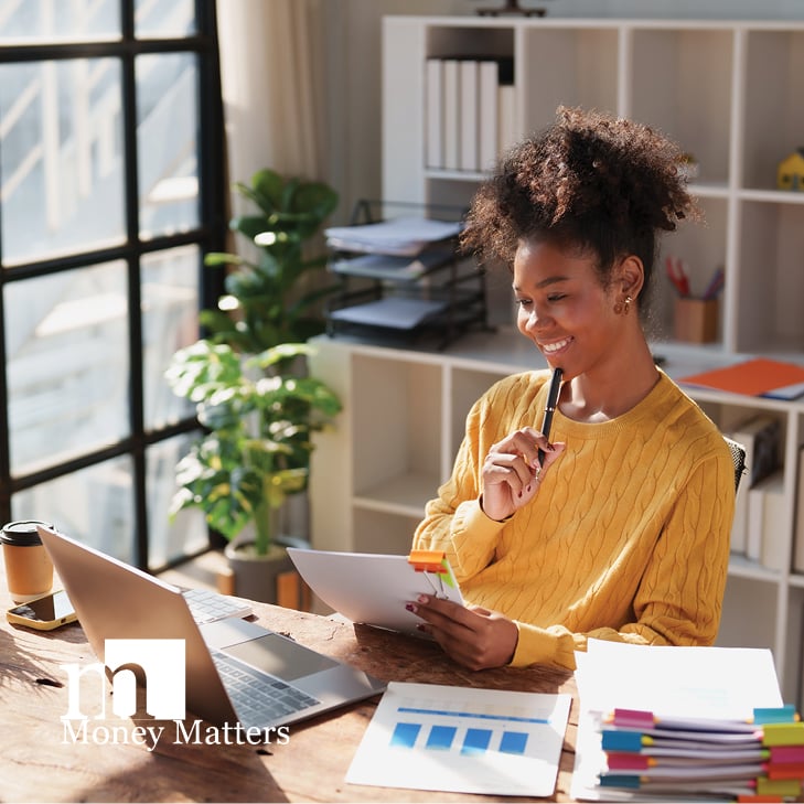 A woman sits at a desk, looking at a laptop.