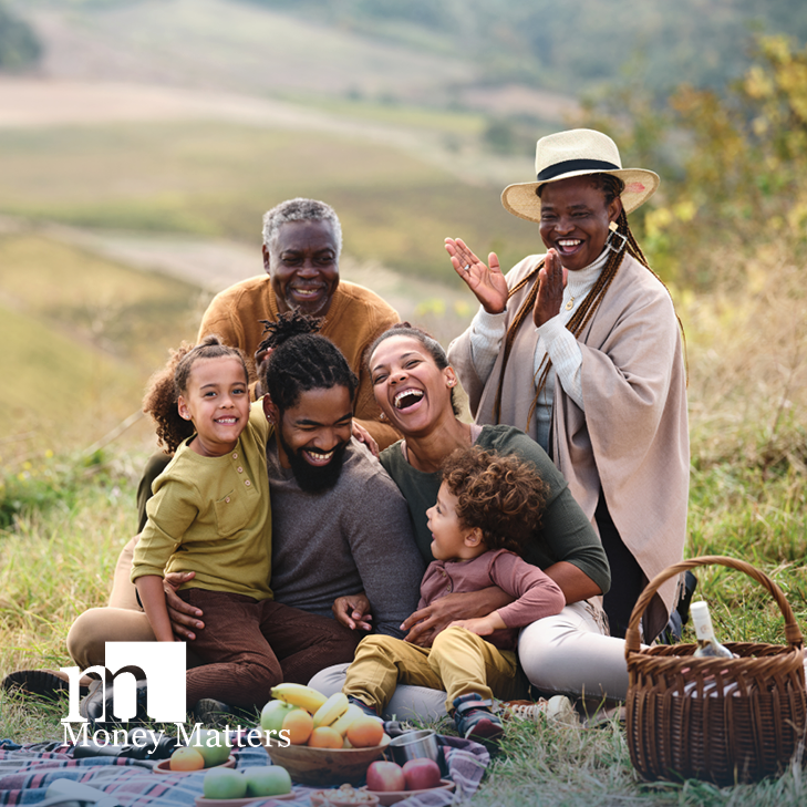 A younger man and woman with two children, and an older man and woman, sit on a blanket in a field.
