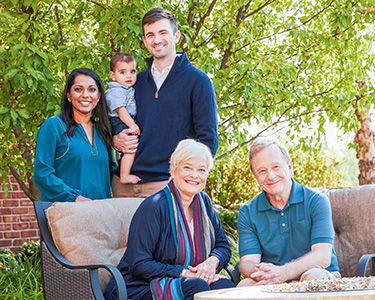 Multi-generational family of five, smiling grandparents, adult children and a young grandchild gathered around an outdoor fire pit.